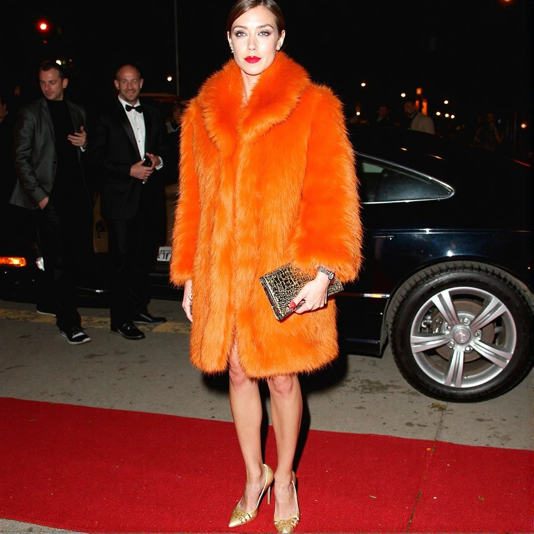 Photograph of a glamorous ensemble with a bright orange faux fur coat, gold heels, and a metallic clutch, standing on a red carpet at night.